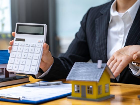Equity - Real estate agent holding up calculator and gesturing to a model house on their desk