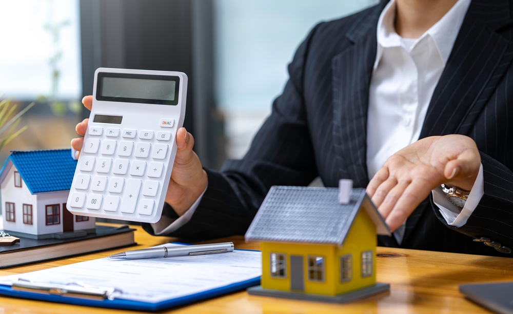 Equity - Real estate agent holding up calculator and gesturing to a model house on their desk