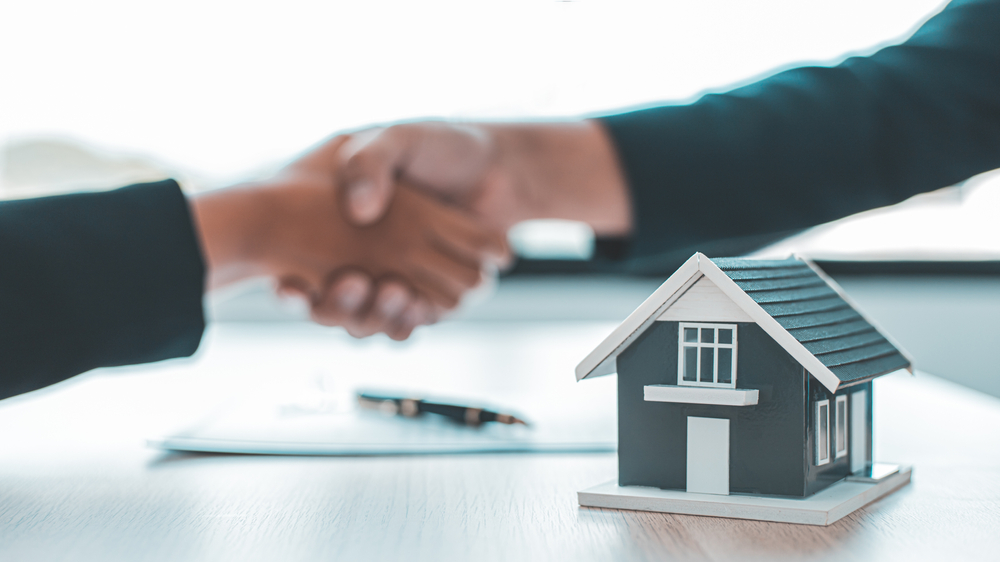 Close up of two people shaking hands in front of a model house, pen, and paperwork