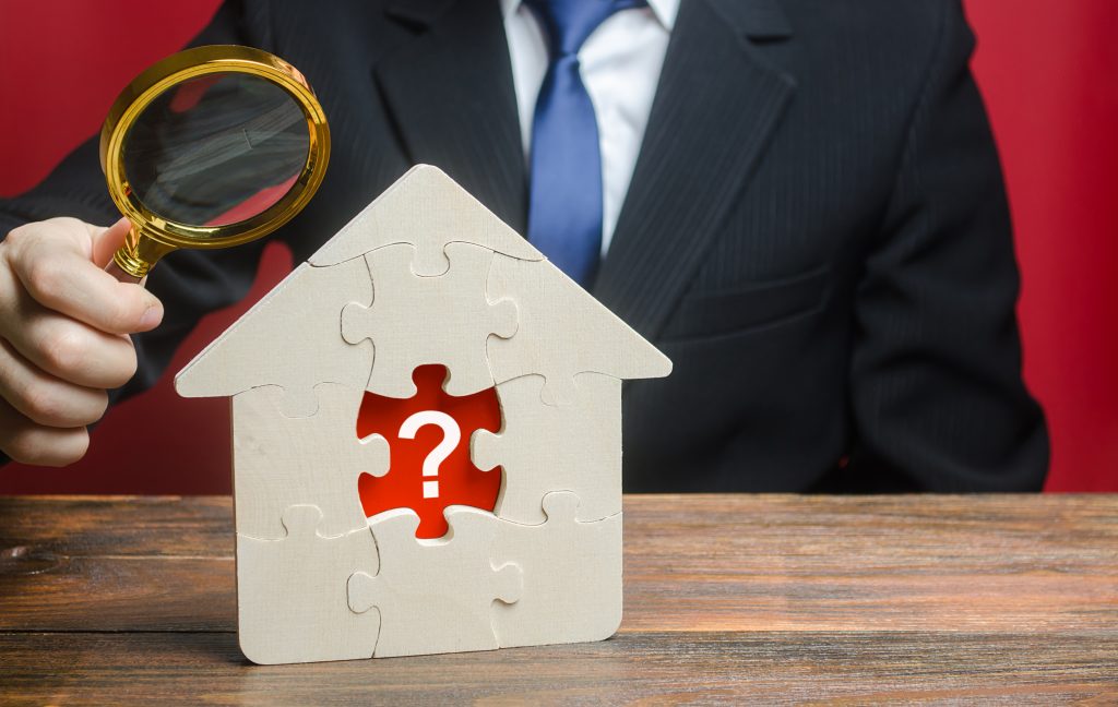 Man pointing a magnifying glass as a house-shaped puzzle with one piece missing in the centre, revealing a question mark