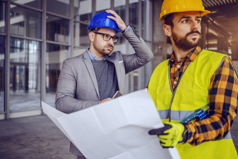 Two upset architects who made mistakes. One holding a map and looking off into the distance. One holding his hard hat